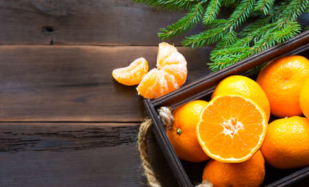 Fresh tangerines in brown box on wooden background with green fir branches. Frame, copy space, holiday and winter fragrance, new year, Christmas. Healthy food, orange juice, shop window. Slice and cutの写真素材
