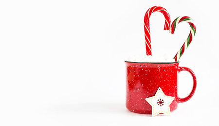Red mug with marshmallows and caramel candy cane in the shape of a heart on a white background and a...