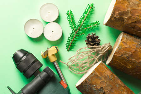 DIY Christmas candle holder made of pine logs, candles, craft rope, fir branches and cones. Layout on a green background. A drill is a tool for manufacturing. Step-by-step instructions, step 1.の写真素材