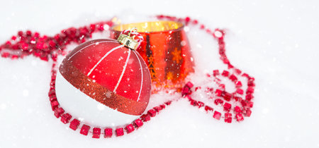 Red Christmas ball on natural white snow with square beads and burning candle in a glass candlestick. Christmas, New year outdoor. Snowfall, festive mood of fairy tales and magic, street decoration.の写真素材