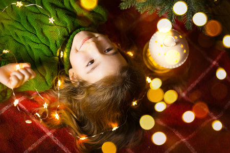 A girl with long hair and garlands lies on a red plaid under a Christmas tree with toys in a warm knitted sweater. Christmas, New Year, children's emotions, joy, expectation of a miracle and giftsの写真素材