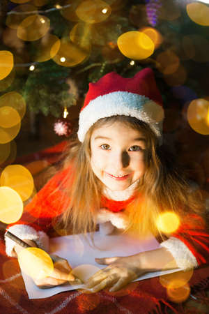 Little girl in a Santa hat and red dress under Christmas tree is dreaming, waiting for the holiday, lying on a plaid blanket. A letter on piece of paper, gifts. New year, Christmas. defocus lightsの写真素材