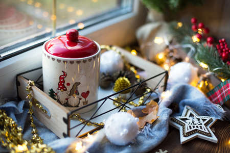 Christmas decor by the window on a cozy wooden windowsill with a mug with a drink and a cookie jar. Fairy lights, Christmas tree branch, breakfast tray still lifeの写真素材