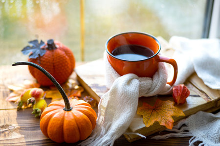 An orange mug in a scarf with hot tea, pumpkins, yellow dry maple leaves, a book on the windowsill,...