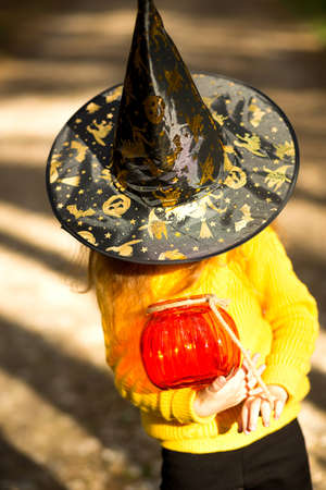 A girl in a witch's hat, in a yellow sweater and with a pumpkin a lantern and a bucket for sweets in autumn forest. Halloween Holiday. Portrait, close-upの写真素材