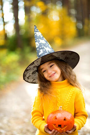 A girl in a witch's hat, in a yellow sweater and with a pumpkin with carved eyes and a mouth Jack o Lantern in a yellow autumn forest. Halloween holidayの写真素材