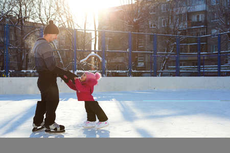 Dad teaches his little daughter to ice skate on a skating rink in the courtyard of multi-storey buildings in the city. Frosty winter sunny day, active winter sports and lifestyleの写真素材