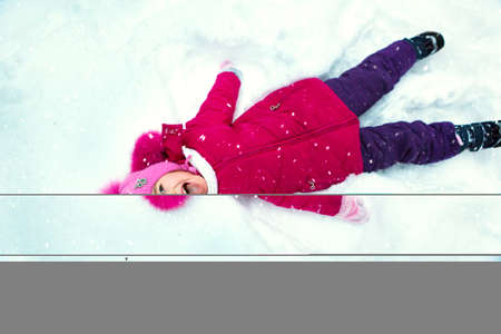 Girl lies on the snow, makes a snow angel, laughs and catches snowflakes with her mouth. Winter, walk the baby in the open air, snow. Warm clothing, knitted hat, fur pompom and hood. Snowball fightの写真素材
