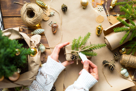 Hands in a white sweater pack a gift for Christmas and new year in eco-friendly materials: kraft paper, live fir branches, cones, twine. Tags with mock up, natural decor, hand made, DIY. Festive moodの写真素材