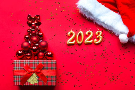 Christmas tree made of glass balls and the numbers 2023 from candles on a red background with gold...