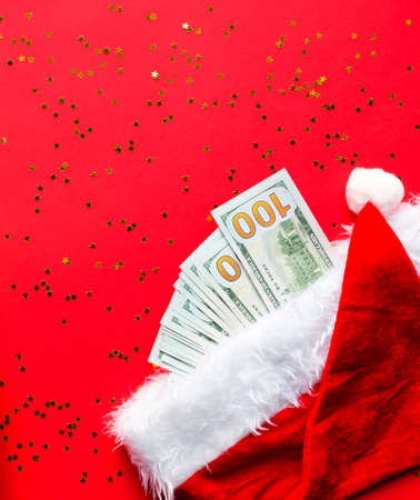 Wad of 100 dollar bills in Santa's hat on a red background with gold sequins. Christmas gift of finance, savings, wealth, expenses in new year. Flatly. Investments, business, money present. Stack $ 100の写真素材