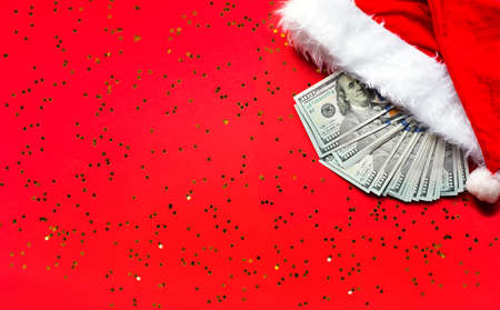 Wad of 100 dollar bills in Santa's hat on a red background with gold sequins. Christmas gift of finance, savings, wealth, expenses in new year. Flatly. Investments, business, money present. Stack $ 100の写真素材