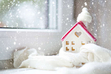 Cozy house is wrapped in a hat and scarf in a snowstorm -window sill decor. Winter, snow - home insulation, protection from cold and bad weather, room heating system. Festive mood, Christmas, New Yearの写真素材