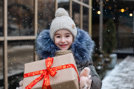 Portrait of joyful girl with a gift box for Christmas on a city street in winter with snow on a festive market with decorations and lights. Warm clothes, knitted hat, scarf and fur. Copy spaceの写真素材