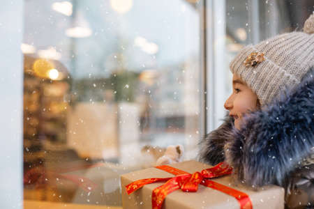 Portrait of joyful girl with a gift box for Christmas near the glass shop window in winter with snow on festive market with decorations and lights. Warm clothes, knitted hat, scarf and fur. Copy spaceの写真素材