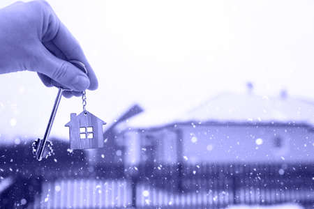 Hand with a Key of the house in winter in the snow. Background of fence and cottage. Give a gift for New Year, Christmas. Moving to new house, mortgage, rent and purchase real estate. Open the door.の写真素材