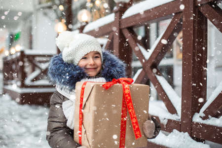 Portrait of joyful girl with a gift box for Christmas on a city street in winter with snow on a festive market with decorations and fairy lights. Warm clothes, knitted hat, scarf and fur. New yearの写真素材