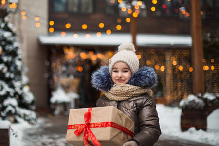 Portrait of joyful girl with a gift box for Christmas on a city street in winter with snow on a festive market with decorations and fairy lights. Warm clothes, knitted hat, scarf and fur. New yearの写真素材