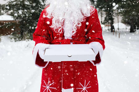 White pizza box in the hands of Santa Claus in white mittens, with a beard, in a red coat. Christmas fast food delivery. New year's eve promotion. Work on public holidays catering. Copy space, mock upの写真素材