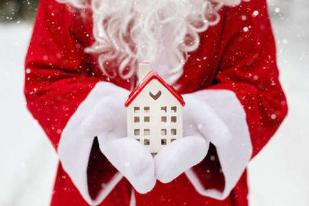 House, miniature cottage in hands of Santa Claus outdoor in snow. Deal for real estate, purchase, construction, relocation, mortgage. Cozy home, Delivery of gifts. Merry Christmas, new year bookingの写真素材