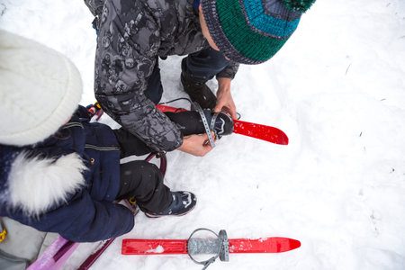 Dad helps his little daughter to put on children's plastic skis. Sports training, skiing, support...