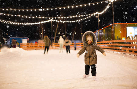 Little girl is skating on ice skates on skating rink in evening, decorated with fairy lights, Christmas trees and fir. Festive mood, Christmas, New Year, holidays, active winter sports and lifestyleの写真素材