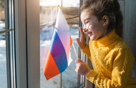 A joyful girl in a yellow sweater with the flag of Russia looks out the window. Patriot, world peace, Victory Day, support and securityの写真素材