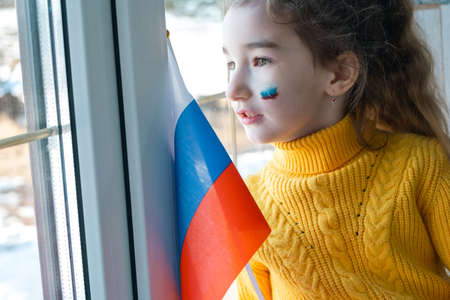 A sad child at the window with the flag of Russia, worries with tears in his eyes. Conflict between Russia and Ukraine, fearの写真素材