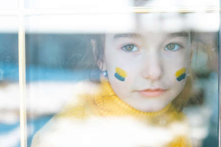 A sad child look at the window with the flag of Ukraine painted on the cheek, worries and fear. Humanitarian aid to children, world peace, security.の写真素材