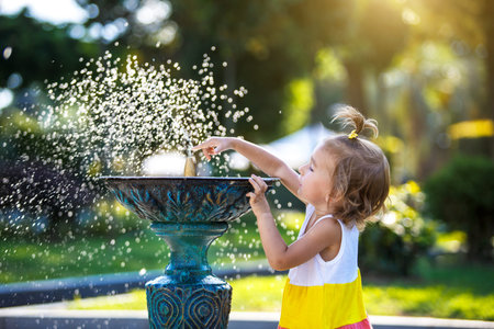 A funny little girl is playing with the spray of a drinking water fountain in the park. Childhood, child, summertime, heat. solar protection for children's skin,の写真素材