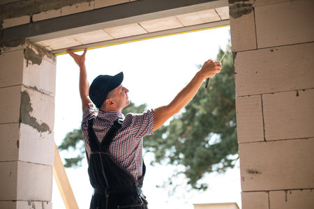 Construction worker at construction site measures the length of window opening and brick wall with tape measure. Cottage are made of porous concrete blocks, work clothes - jumpsuit and baseball capの写真素材