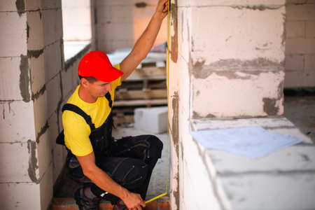 Construction worker at construction site measures the length of window opening and brick wall with tape measure. Cottage are made of porous concrete blocks, work clothes - jumpsuit and baseball capの写真素材