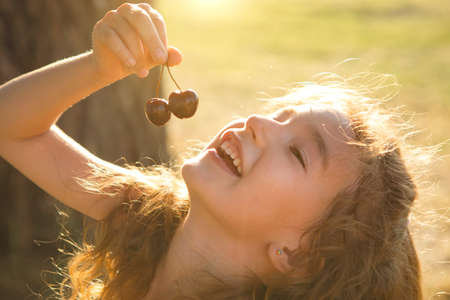 A cheerful sweet girl with cherry berries in her mouth. Funny summer portrait of a child with a cherry, gifts of summer, summer time.の写真素材