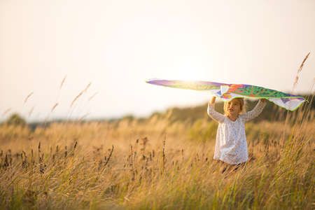 A girl runs into a field with a kite, learns to launch it. Outdoor entertainment in summer, nature and fresh air. Childhood, freedom and carelessness. A child with wings is a dream and a hope.の写真素材