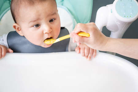 Mom feeds the baby with a spoon of vegetable puree at the children's feeding table. Baby's appetite, healthy nutrition, introduction of complementary foods. copyspace mock upの写真素材