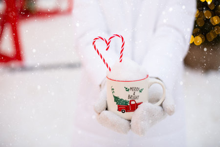 Woman with mug with snow, candy cane and inscription Merry and Bright in her hands outdoor in warm...