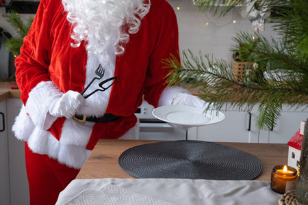 Santa Claus sets the table at home in the white kitchen, puts a plate, puts cutlery fork and knife. Preparing for the celebration of Christmas, setting the table for guestsの写真素材