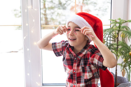 Girl in a Santa hat sits on the windowsill of a house near the Christmas tree and puts Candy cane to her eyes like glasses. Child is having fun and making faces, waiting for Christmas and New yearの写真素材