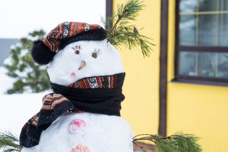 Funny snowman in a hat and scarf on the background of a yellow house in the yard. Winter, winter entertainment, snowfallの写真素材