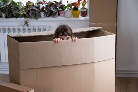 The girl hides in a big box and looks out of it funny in a room inside the home. Moving to a new...