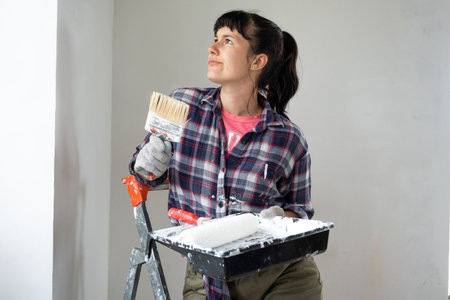 puzzled woman in paint roller and white paint for walls in hands close-up portrait. Construction work and cosmetic repairs in house, wall painting, tinting, finishing work with your own handsの写真素材