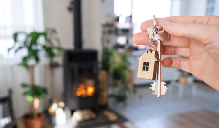Key to house with keychain against background of fireplace stove with fire and firewood. Cozy home hearth. Building, design, project, moving to new house, mortgage, rent and purchase real estateの写真素材