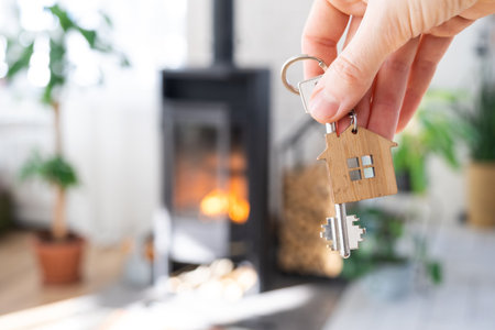 Key to house with keychain against background of fireplace stove with fire and firewood. Cozy home hearth. Building, design, project, moving to new house, mortgage, rent and purchase real estateの写真素材