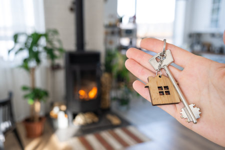 Key to house with keychain against background of fireplace stove with fire and firewood. Cozy home hearth. Building, design, project, moving to new house, mortgage, rent and purchase real estateの写真素材