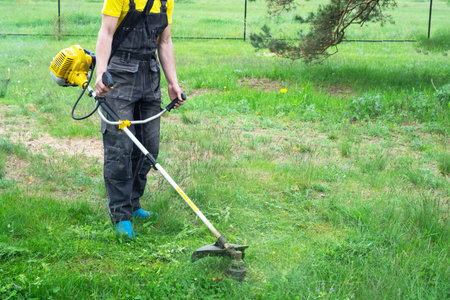 A male gardener mows the green grass of the lawn in the backyard with a gasoline mower. Trimmer for...