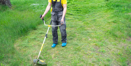 A male gardener mows the green grass of the lawn in the backyard with a gasoline mower. Trimmer for...