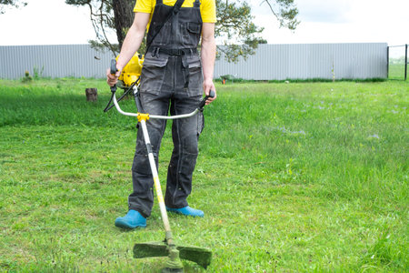 A male gardener mows the green grass of the lawn in the backyard with a gasoline mower. Trimmer for...