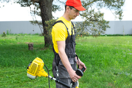 A male gardener mows the green grass of the lawn in the backyard with a gasoline mower. Trimmer for...