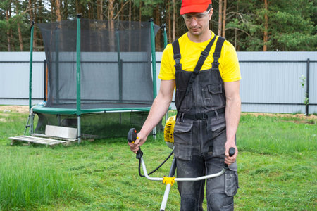 A male gardener mows the green grass of the lawn in the backyard with a gasoline mower. Trimmer for...