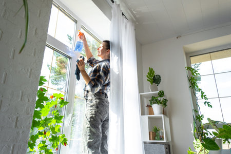 Woman manually washes the window of the house with a rag with spray cleaner and mop inside the...